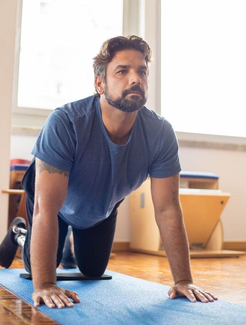 A determined man with a prosthetic leg kneels on a yoga mat for pilates in a bright room.